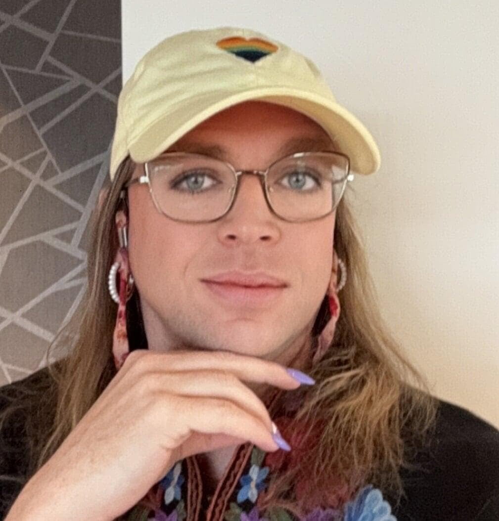 An image of Deanna Grey. She's wearing a yellow hat with a rainbow heart in the center. Her hair is long and drapes along her shoulders. Her hand is resting under her chin. Her fingernails are long and painted purple.